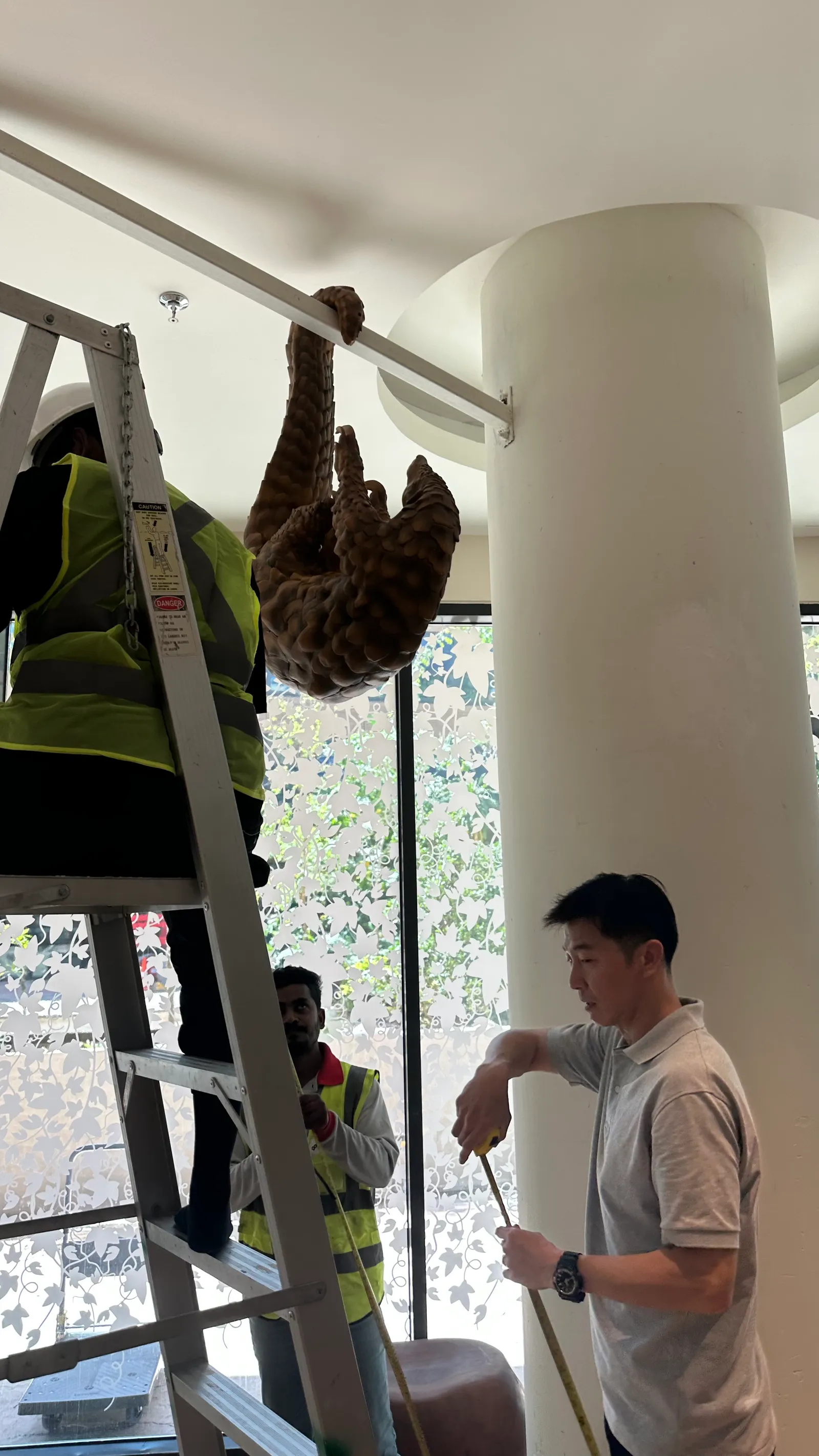 A worker on a ladder fitting a pangolin figure to the branch beside the white trunk, while a colleague below holds a pole