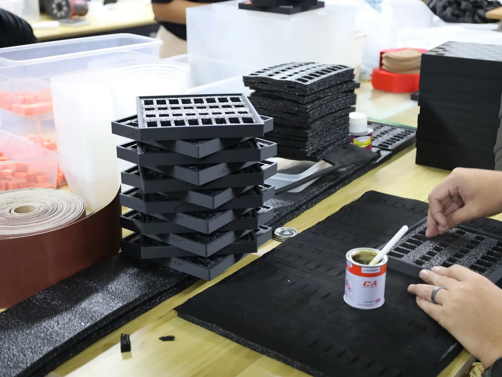 A team member gluing a laser-cut foam insert into a black tray, a stack of finished trays and foam sheets behind