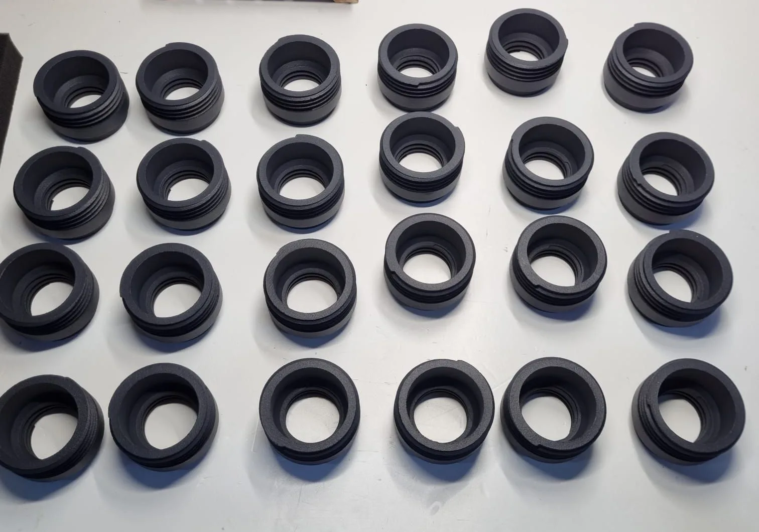 Twenty-four black PA12 nylon bottle attachments set out in four rows on a white bench, each with an outer thread above a smooth band