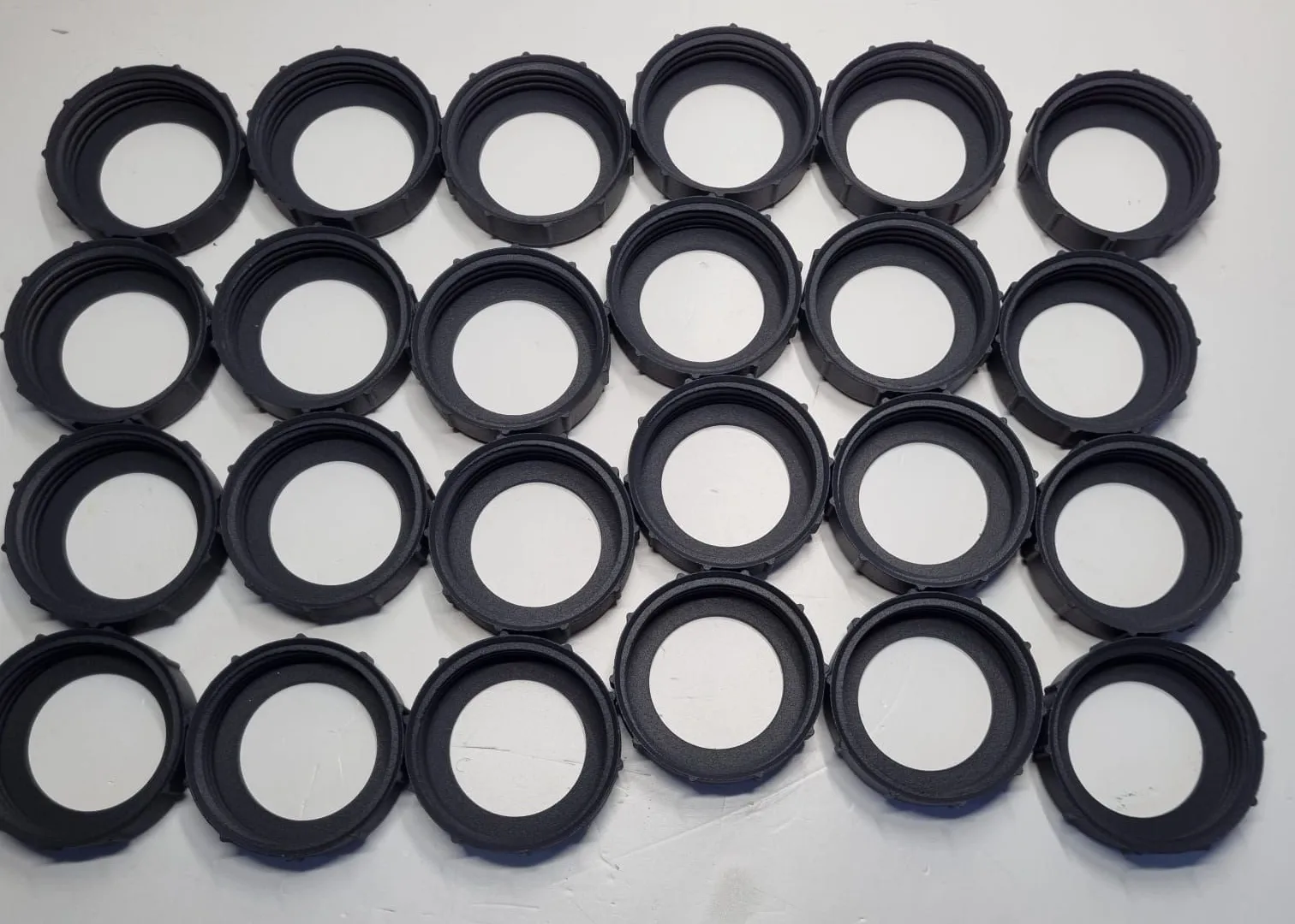 Twenty-four black ring caps in four rows on a white bench, each with a ribbed outer edge and a thread inside