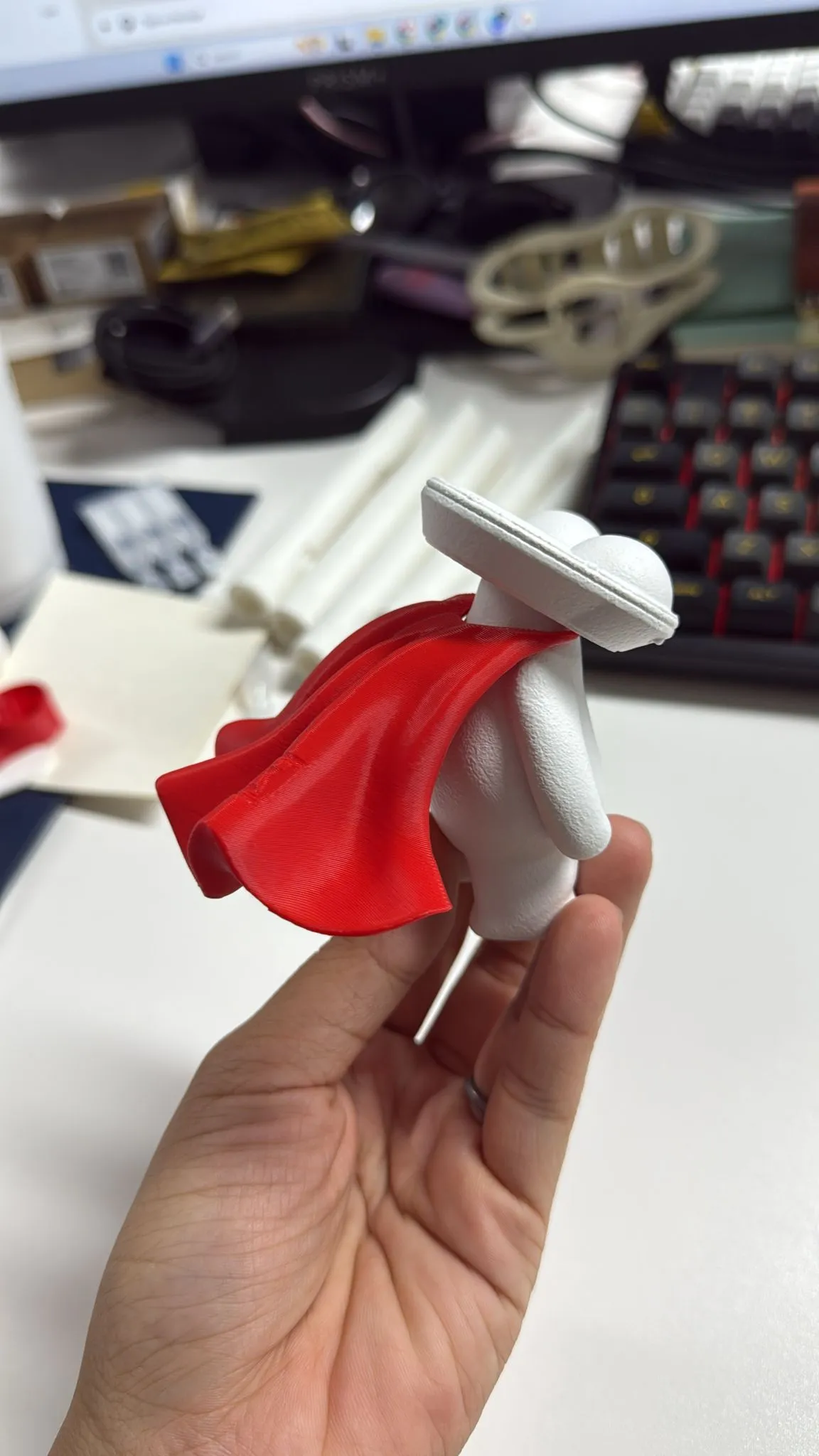 A hand holding a white figure wearing a red test cape, printed to check the fit at the neck
