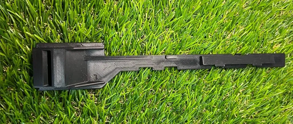 The new 3D printed latch arm in black lying on grass: the square catch at one end, the long notched arm and the raised rail along it