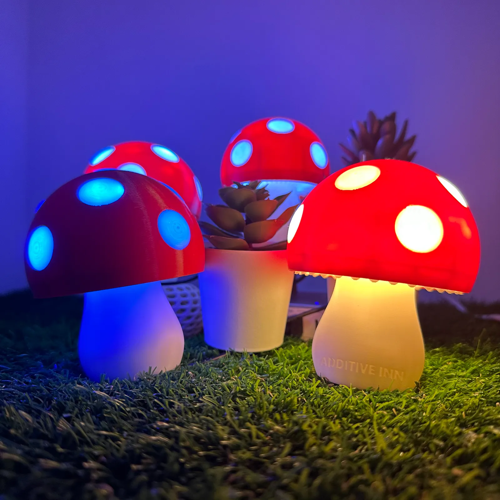 Four finished mushroom lamps lit up on artificial grass in a dim room, their red caps glowing with white and blue spots and the front stem lettered ADDITIVE INN