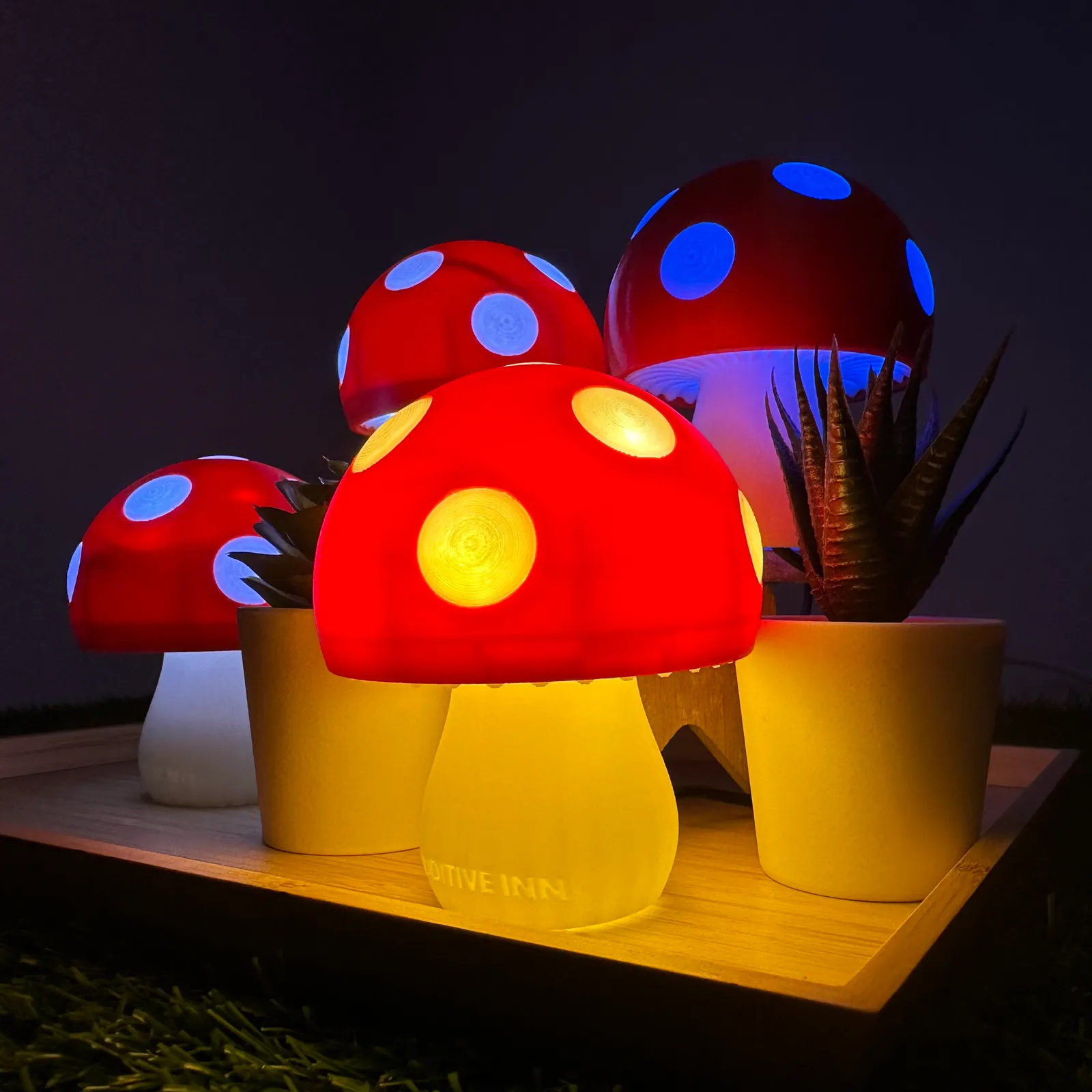 A group of lit mushroom lamps among potted plants on a wooden tray, the front lamp glowing warm yellow and one behind glowing blue