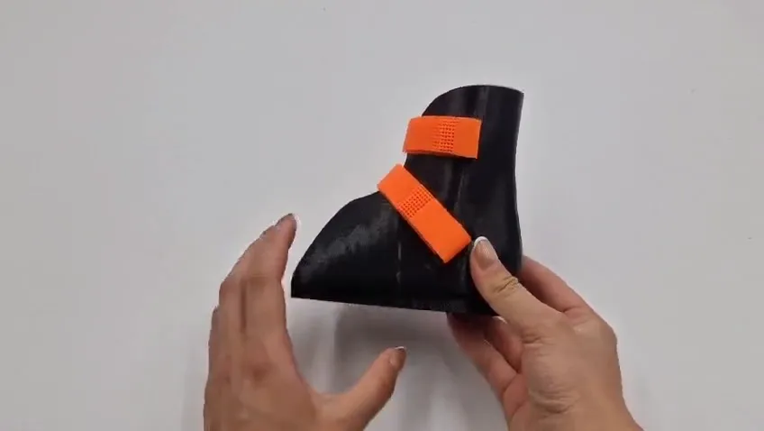 Two hands holding a black 3D printed ankle brace with two orange straps, about to squeeze its flexible shell