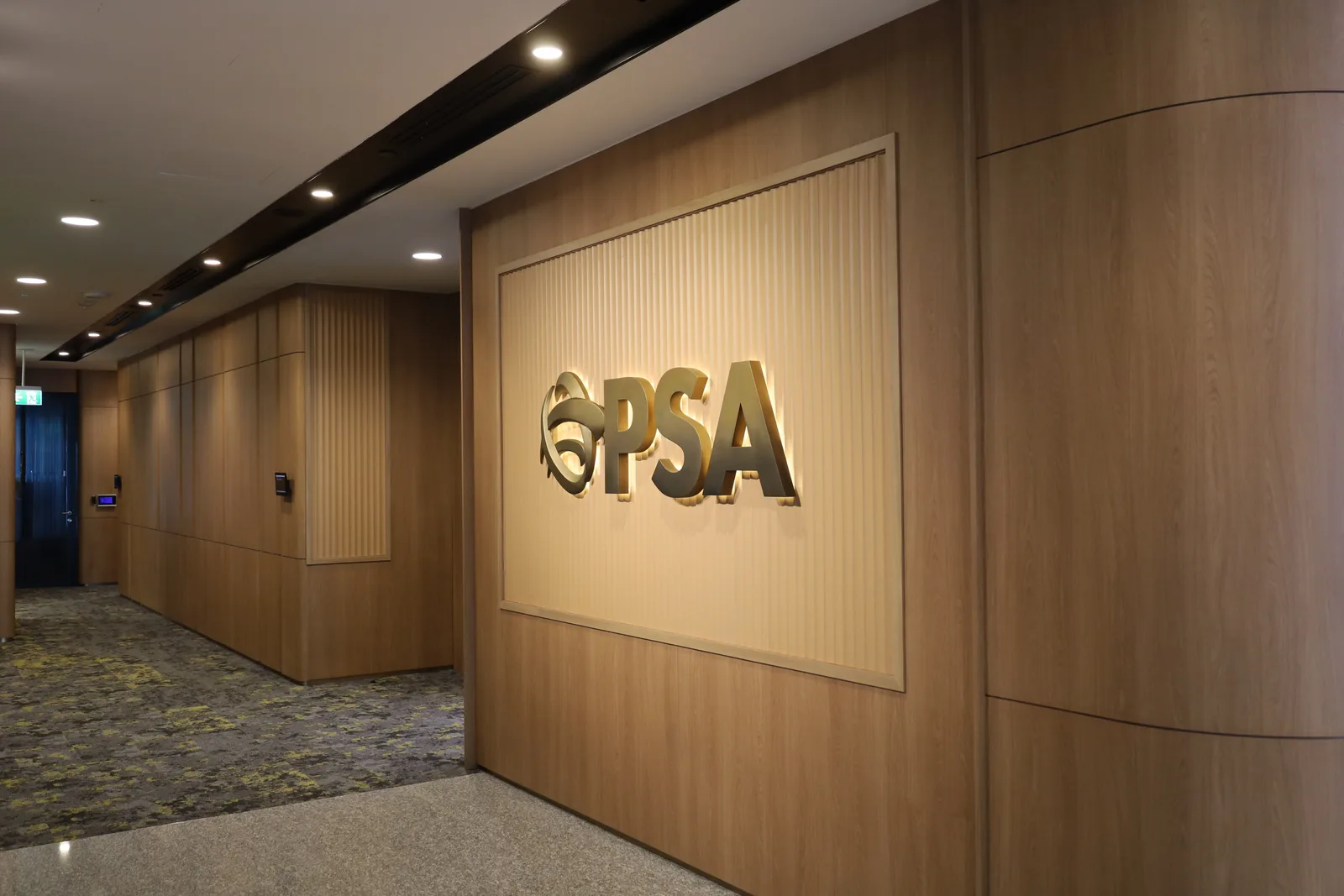 The PSA logo and letters in brushed gold glowing with a warm halo on a fluted wooden feature wall beside an office corridor