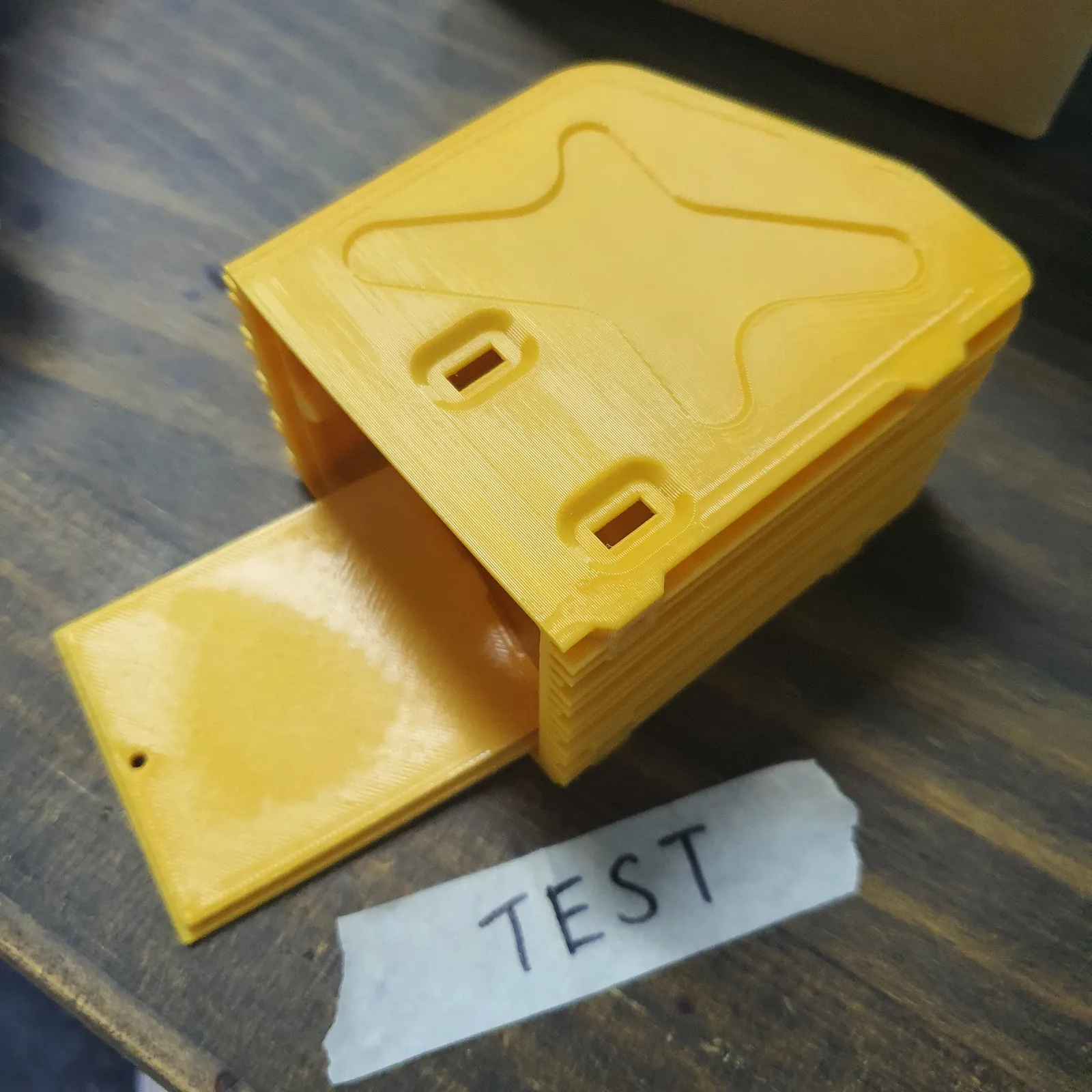 The yellow FDM test print of the enclosure on a workbench, its front panel folded out, with a strip of tape marked TEST