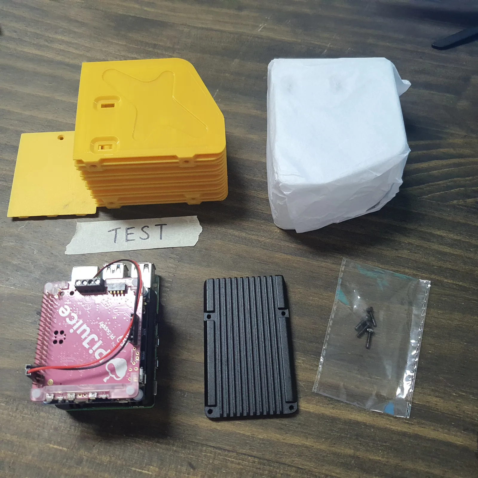 The parts laid out on a workbench: the yellow test shell, the white resin shell still wrapped in tissue, the board stack with its battery board on top, a black heatsink lid and a bag of screws