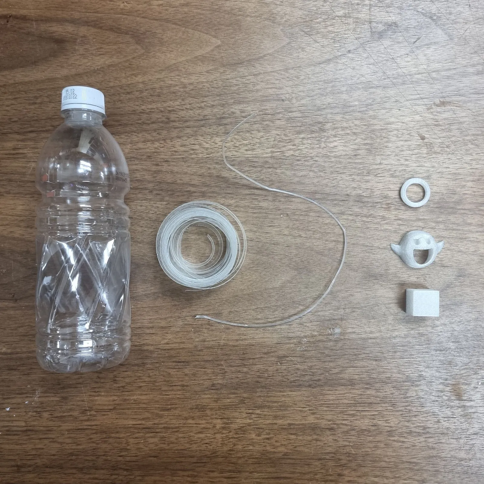 A clear PET drinking bottle on a wooden table beside a coil of cut strip, a length of filament and three small white test prints