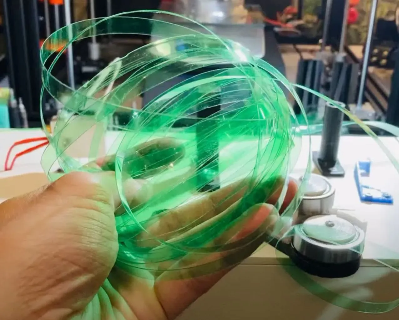 A hand holding a loose coil of green strip cut from a plastic bottle, on a bench in front of a 3D printer