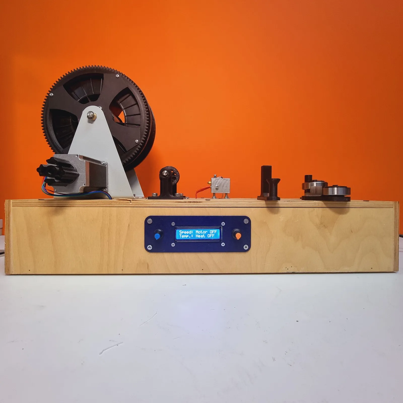 The filament rig head-on against an orange wall: a large geared spool and its motor on the left, the heated nozzle in the middle and the strip feed on the right, on a plywood base