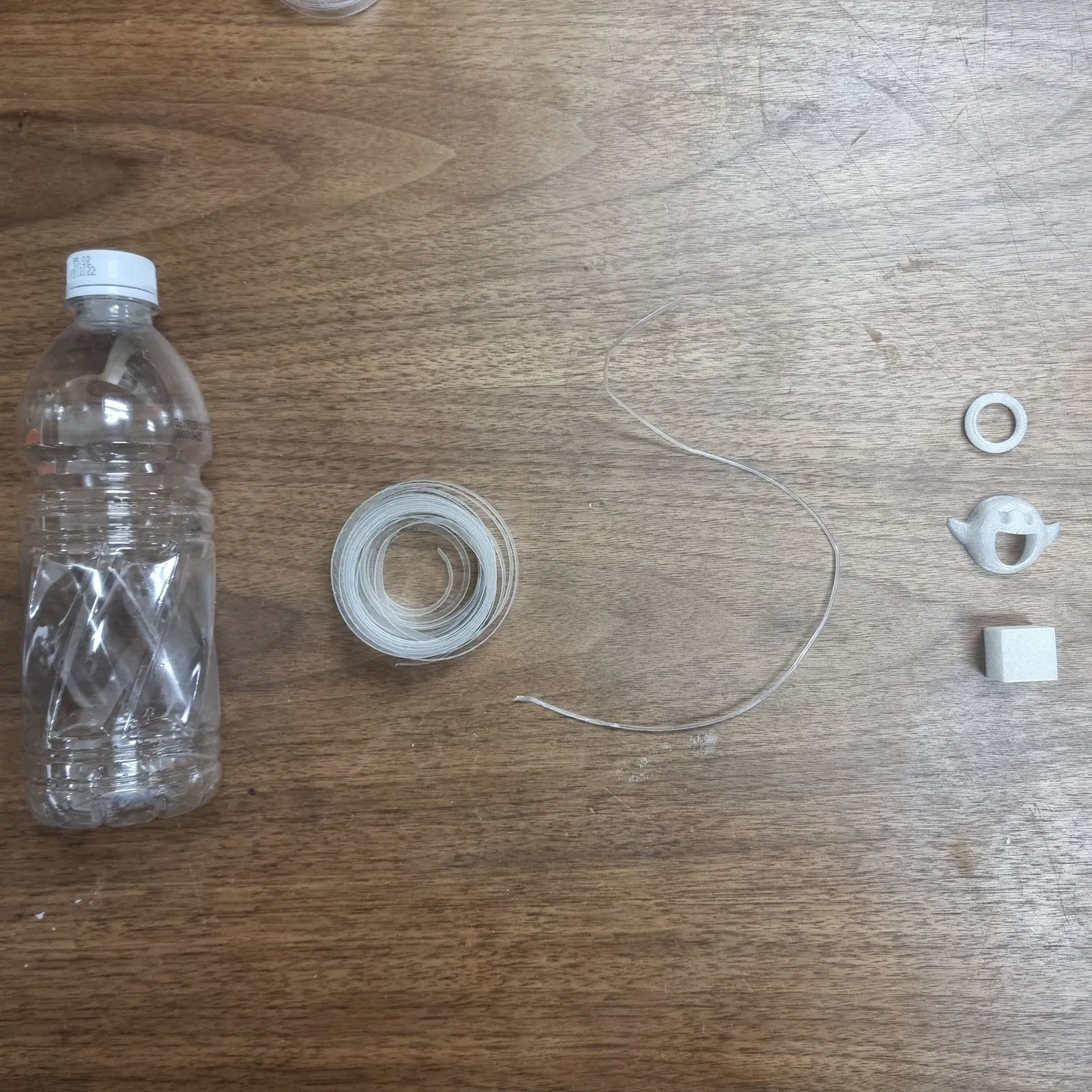Top view of a clear PET bottle, a coil of cut strip, a loop of filament and a ring, a ghost and a cube printed in white, in a row on a wooden table