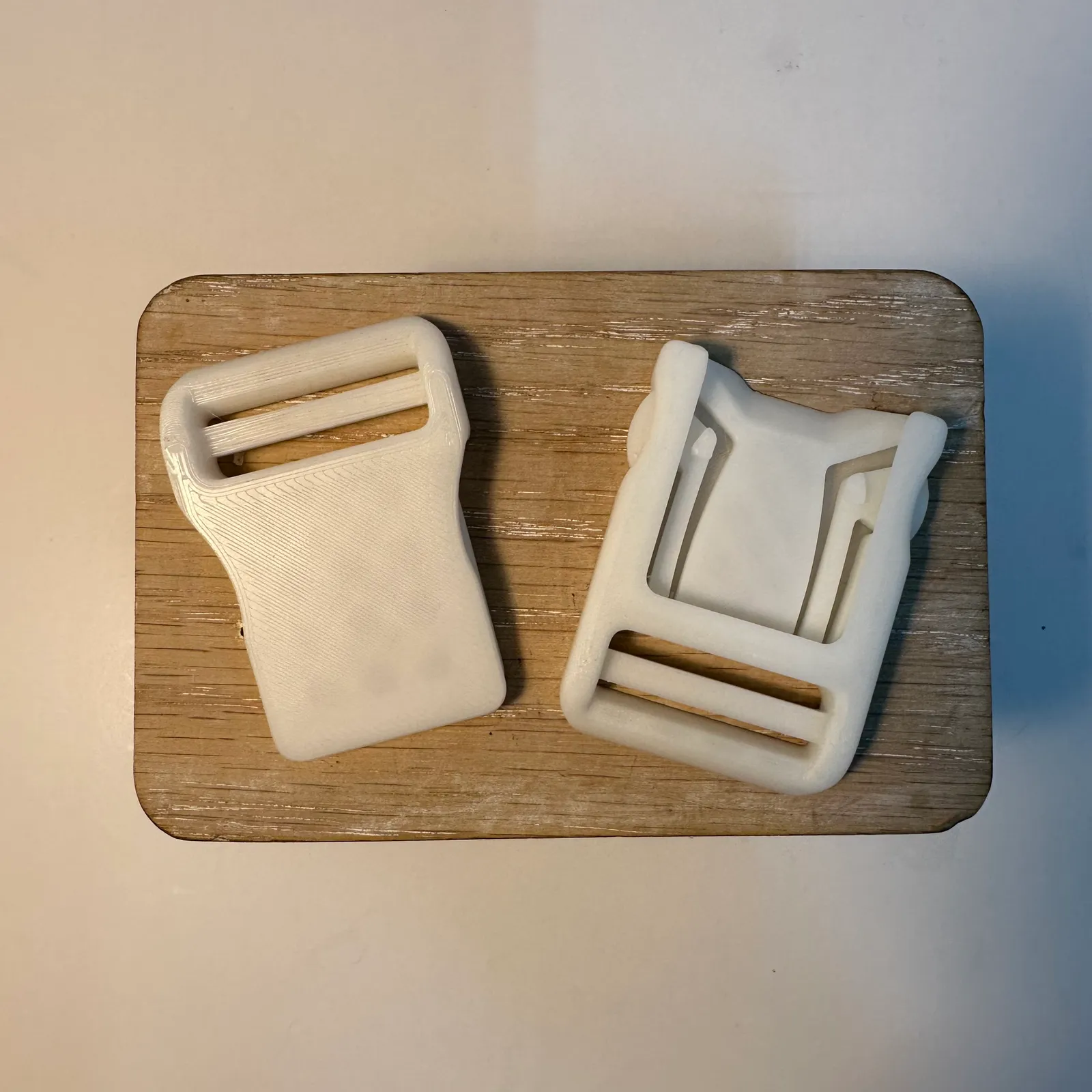 The two halves of the white buckle apart on a wooden stand, seen from above: the socket on the left, and the clip on the right with its two springy arms and strap slot