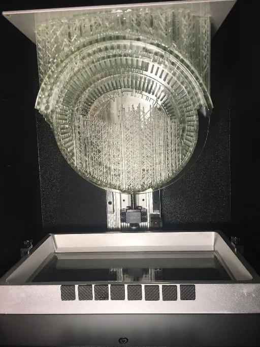 The Colosseum model hanging from the build plate of a resin printer, above the resin tray