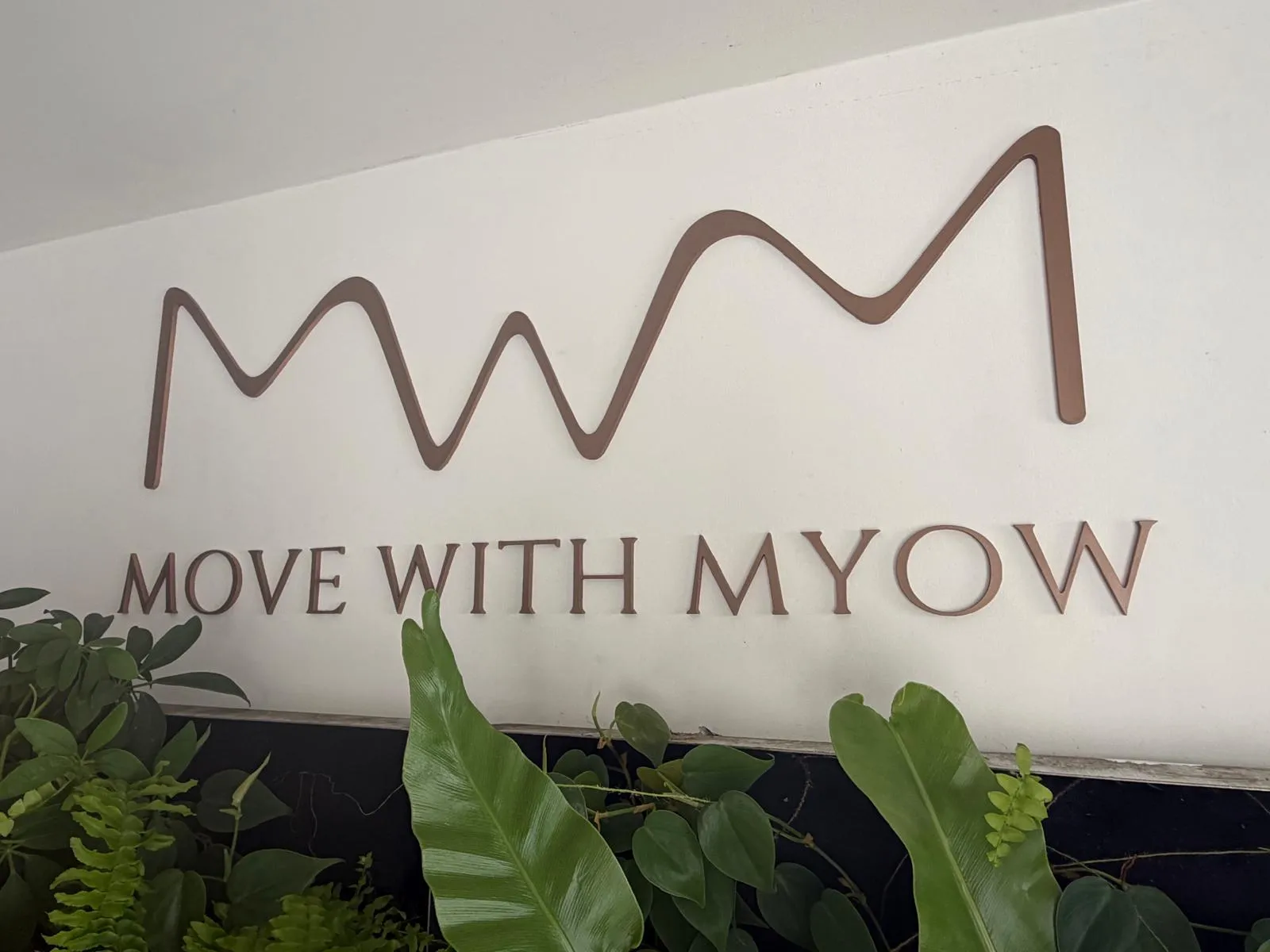 The rose gold Move with Myow sign seen at an angle, each letter standing slightly off the white wall