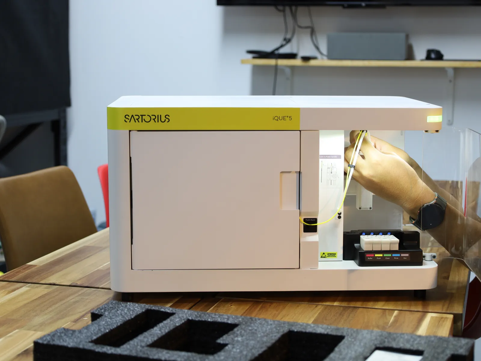 A hand fitting parts into the lit bay of the iQue 5 model, with the cut foam of its travel case on the table in front