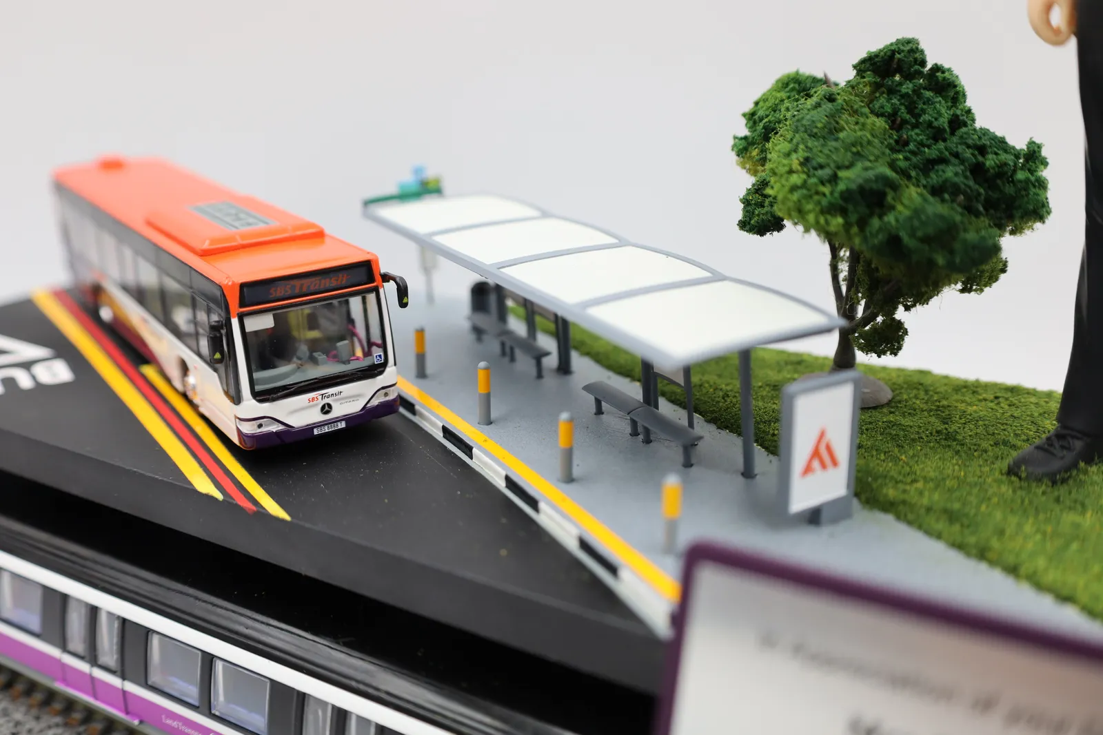 Close-up of the model bus on the road deck beside the bus stop shelter, with a model tree and flocked grass behind