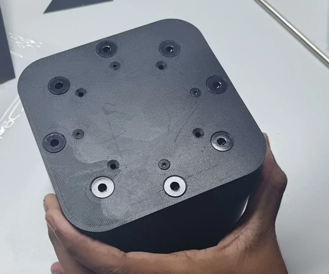 The closed cup mould in a hand, seen from its bolted face: socket head bolts and two steel washers set into the printed plate