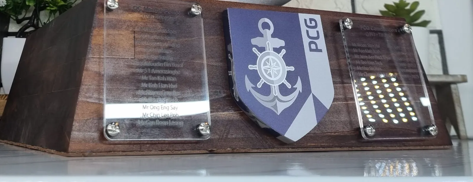 The front of the dark wooden base: a blue and silver PCG anchor shield between two clear acrylic plaques screwed on, listing commanders' names