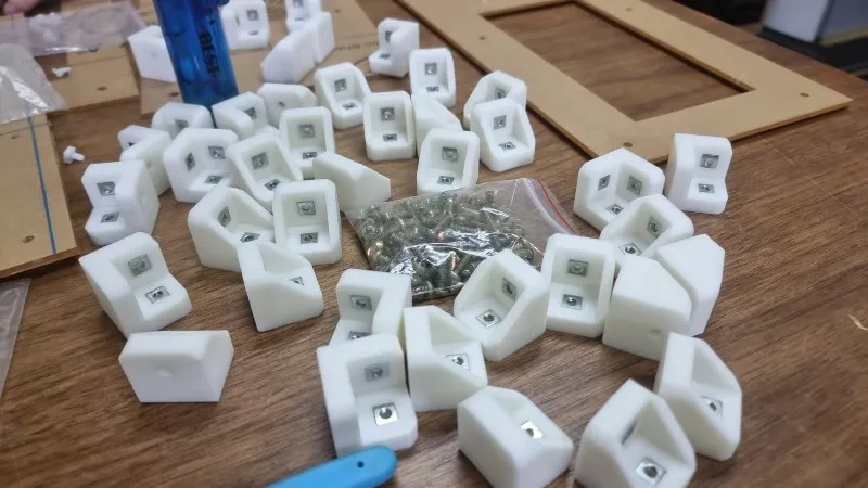 A pile of white tough resin brackets on a table, each with a square nut pocket, beside a bag of screws