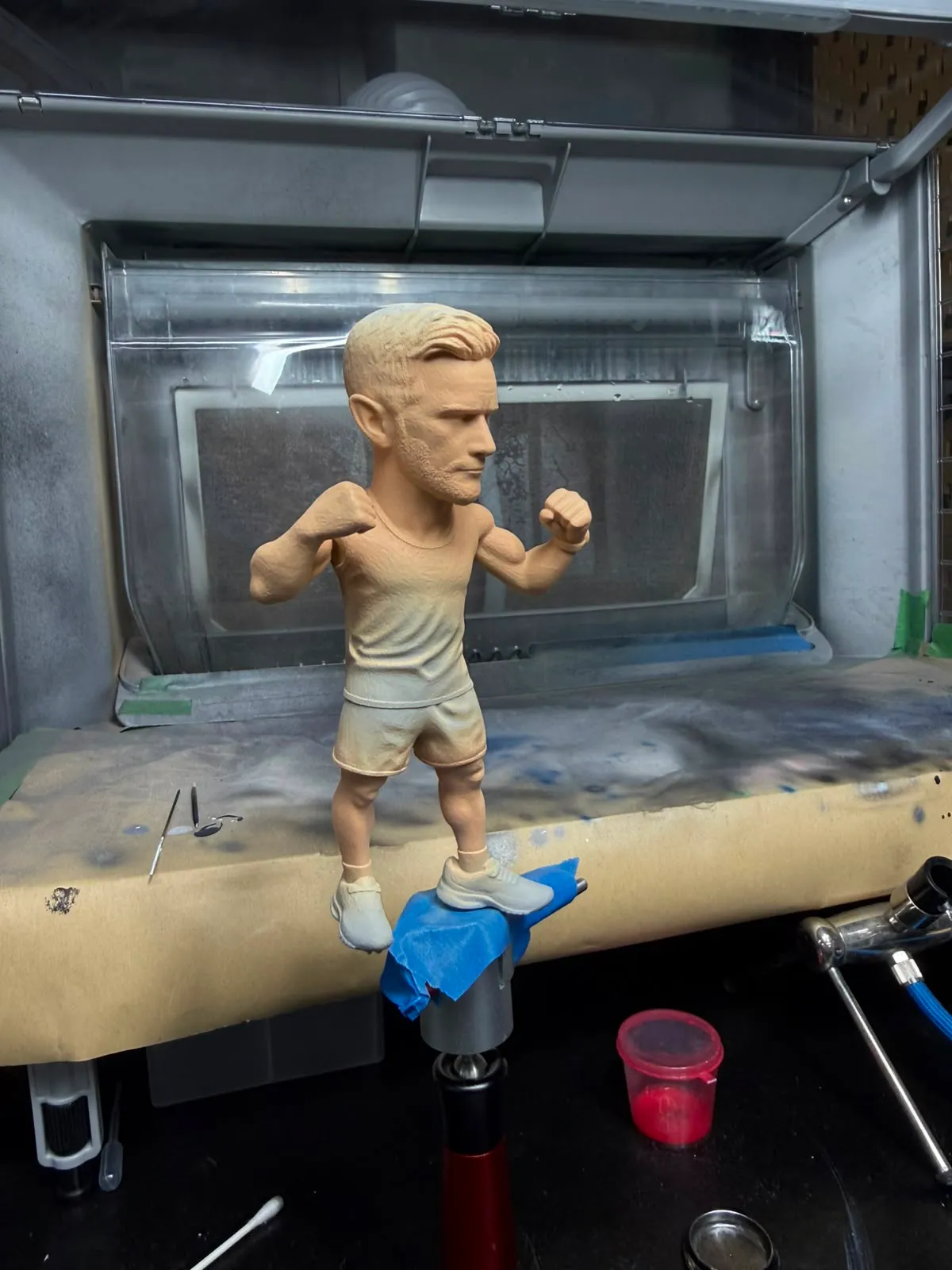 The figure in the spray booth with its first airbrushed skin tones, held on a painting clamp
