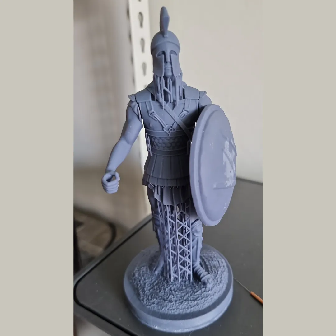 The raw grey resin print of the Spartan with shield, standing on its textured round base, with lattice supports still under the legs