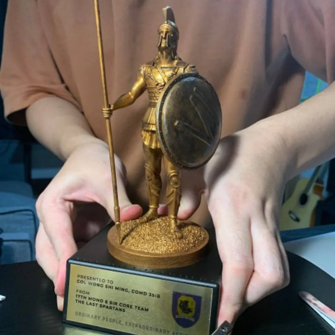 Hands setting the bronze Spartan onto its black base, above the gold plate with the 6 SIR crest