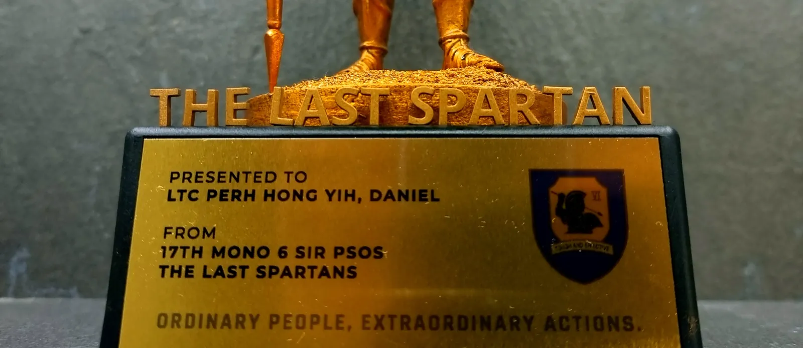 Close-up of the base: raised THE LAST SPARTAN letters above a gold plate presented to LTC Perh Hong Yih, Daniel from 17th Mono 6 SIR PSOs, The Last Spartans, with the 6 SIR crest and the motto Ordinary people, extraordinary actions
