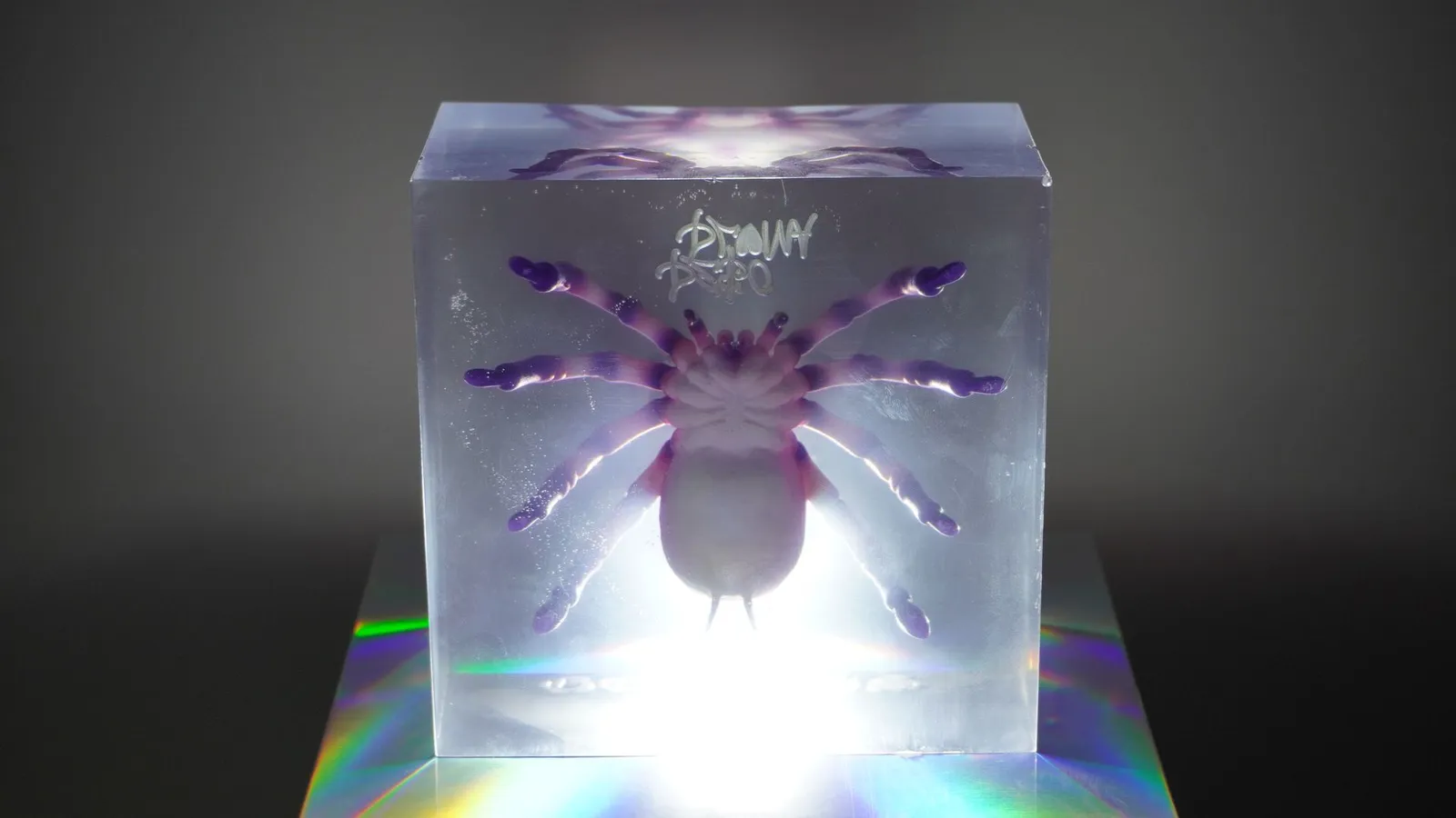 A resin block standing on a holographic base, lit from behind so the clear resin glows around the purple spider