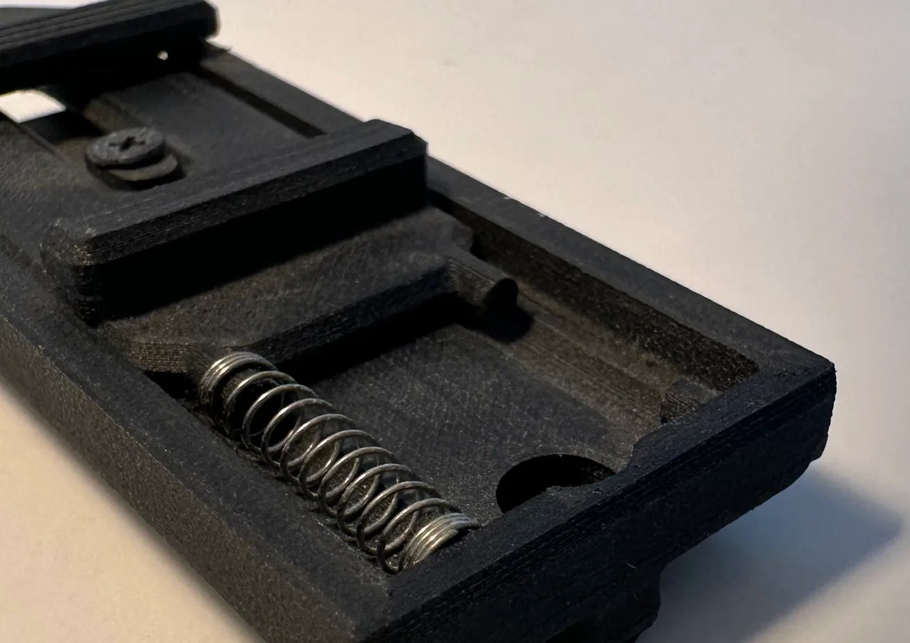 Close-up of the steel coil spring seated in its printed pocket, with the latch grip above it and the screw head beyond