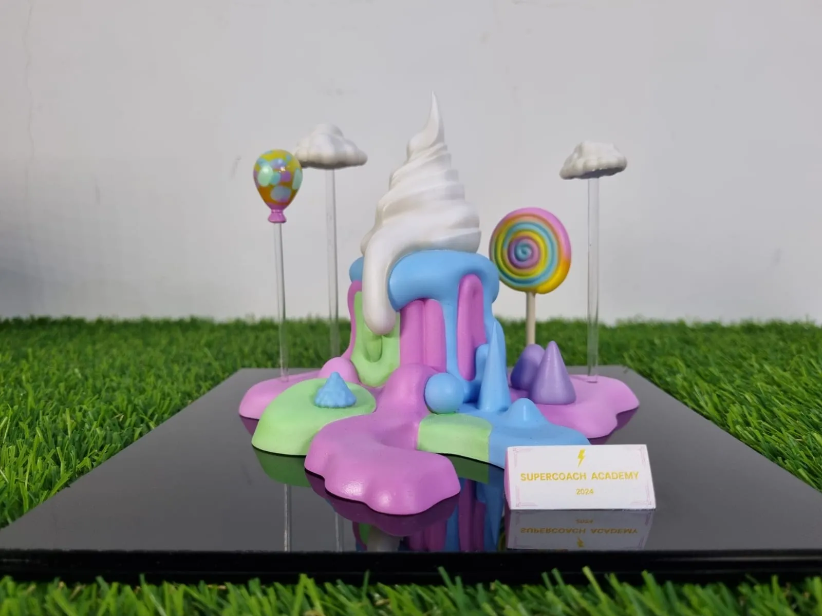 The finished cake trophy: a pearl-white soft-serve swirl melting over pink, mint and blue drips, with a balloon, a lollipop and two clouds on clear rods, on a glossy black base