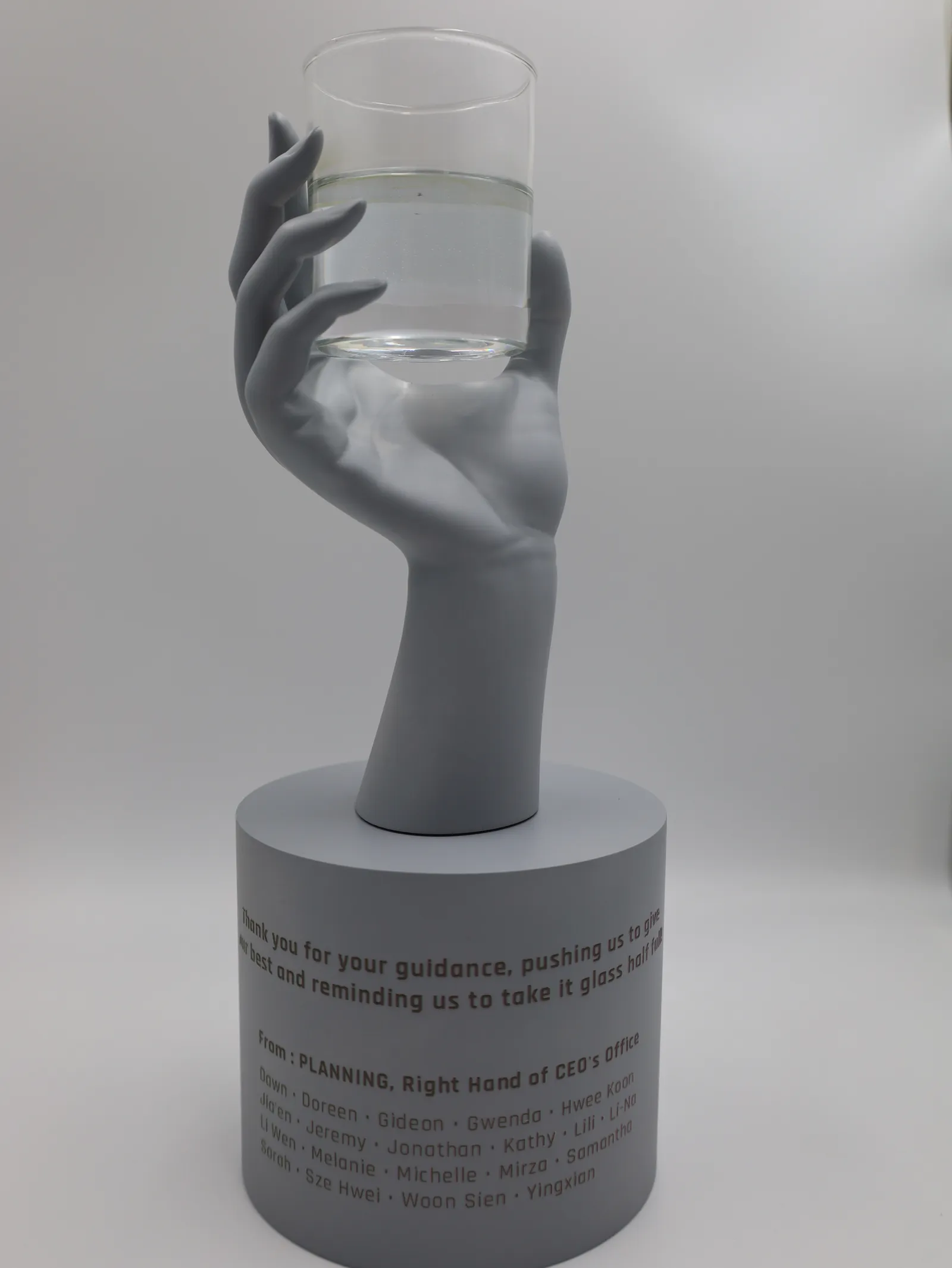 The finished hand trophy from the front: a grey hand holding a clear glass of water above a round base lettered with a message and names