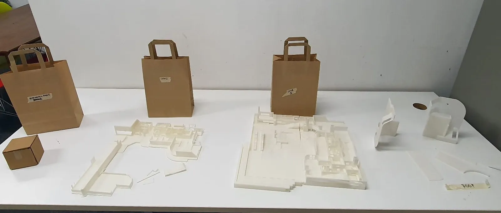 Unpainted printed parts laid out on a table beside paper bags labelled by level, with the roof pieces marked with tape