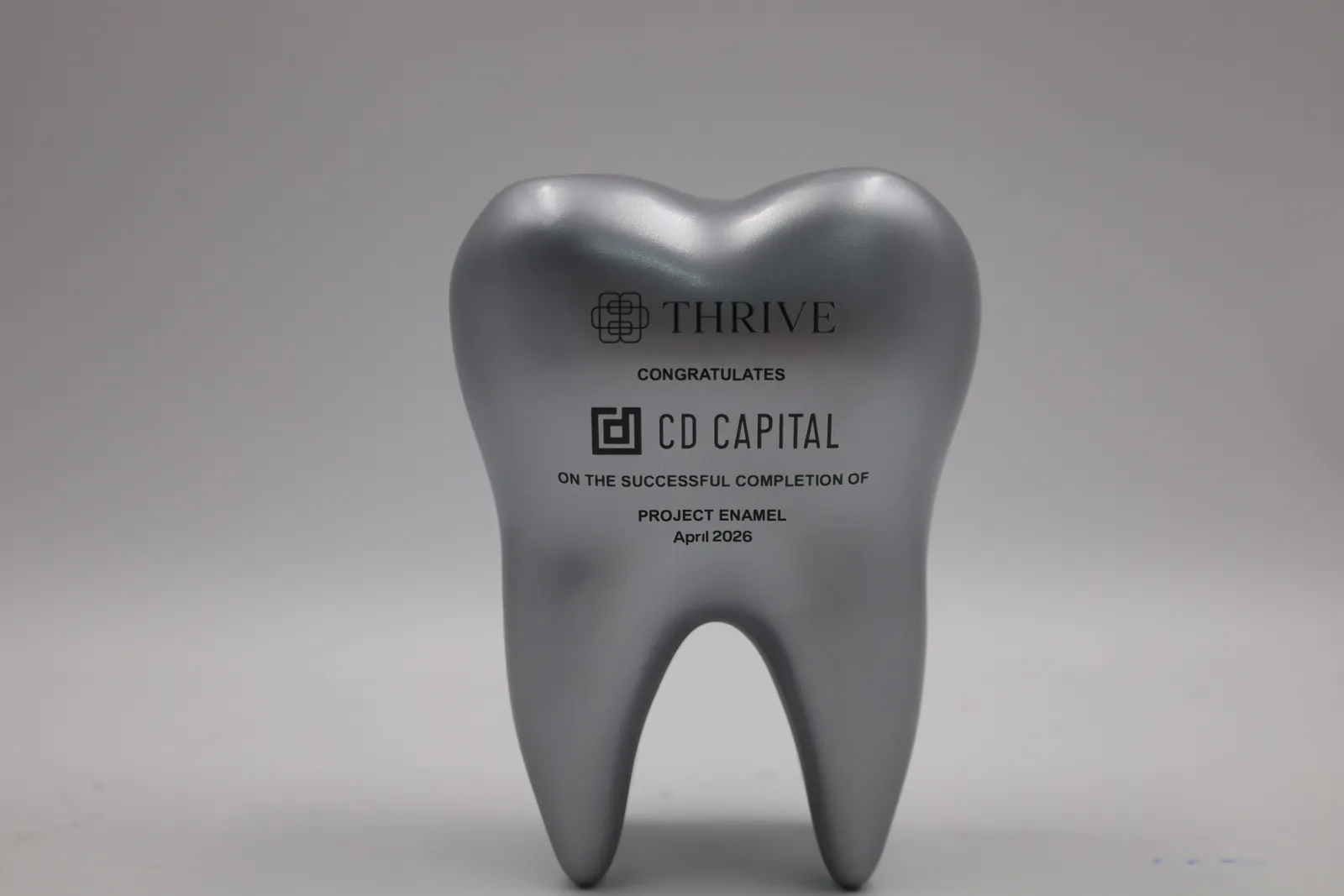 A silver tooth-shaped trophy standing on its roots, with the Thrive and CD Capital logos and a congratulations message printed in black on the front