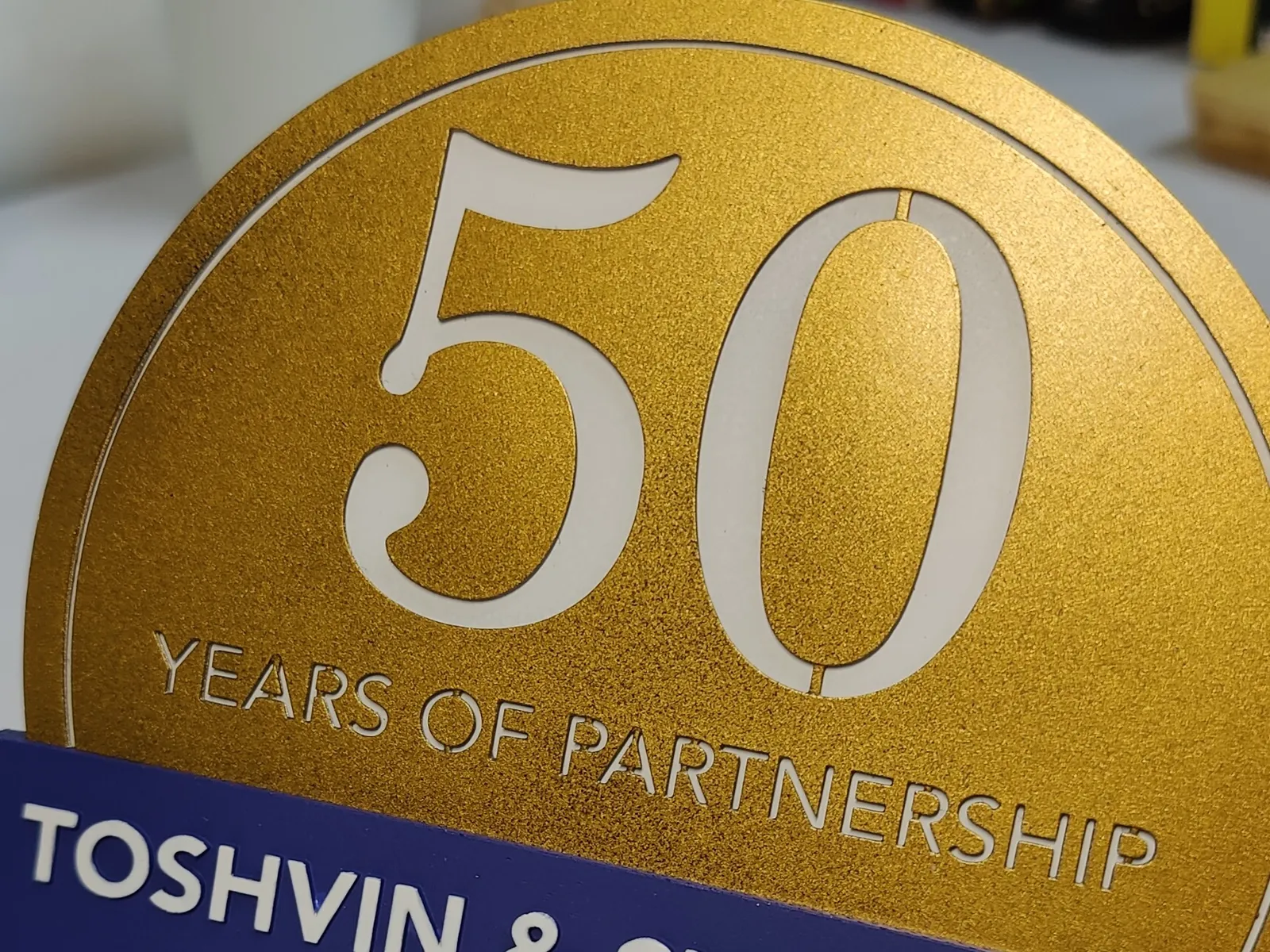 Close-up of the gold coin: a white 50 and YEARS OF PARTNERSHIP cut through the metallic gold, with a thin white ring round the edge