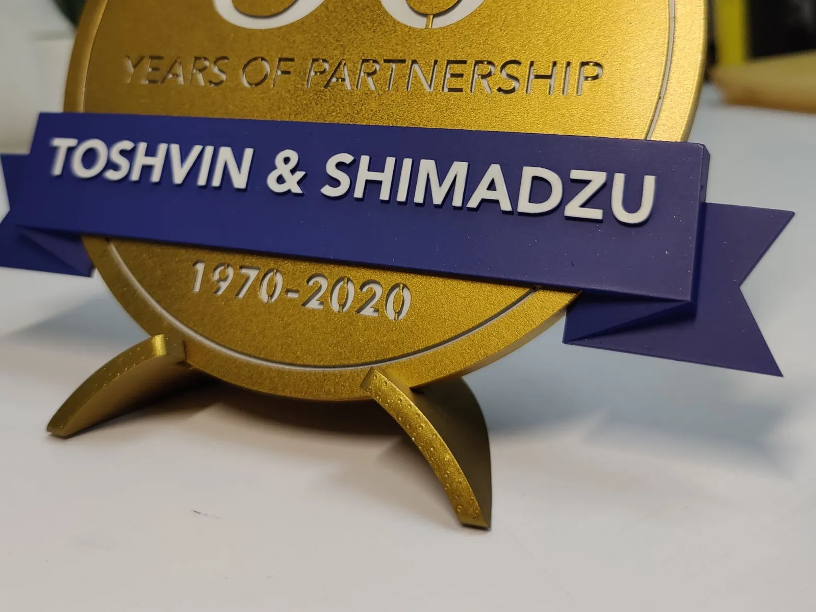 Close-up of the royal-blue banner with raised white TOSHVIN & SHIMADZU lettering, its folded tails, 1970-2020 below and the two curved gold legs