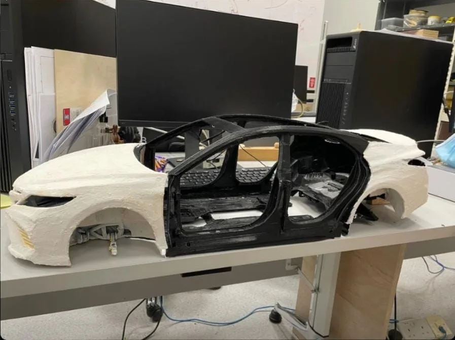 A printed car body shell in white around a black printed inner frame, on a studio bench