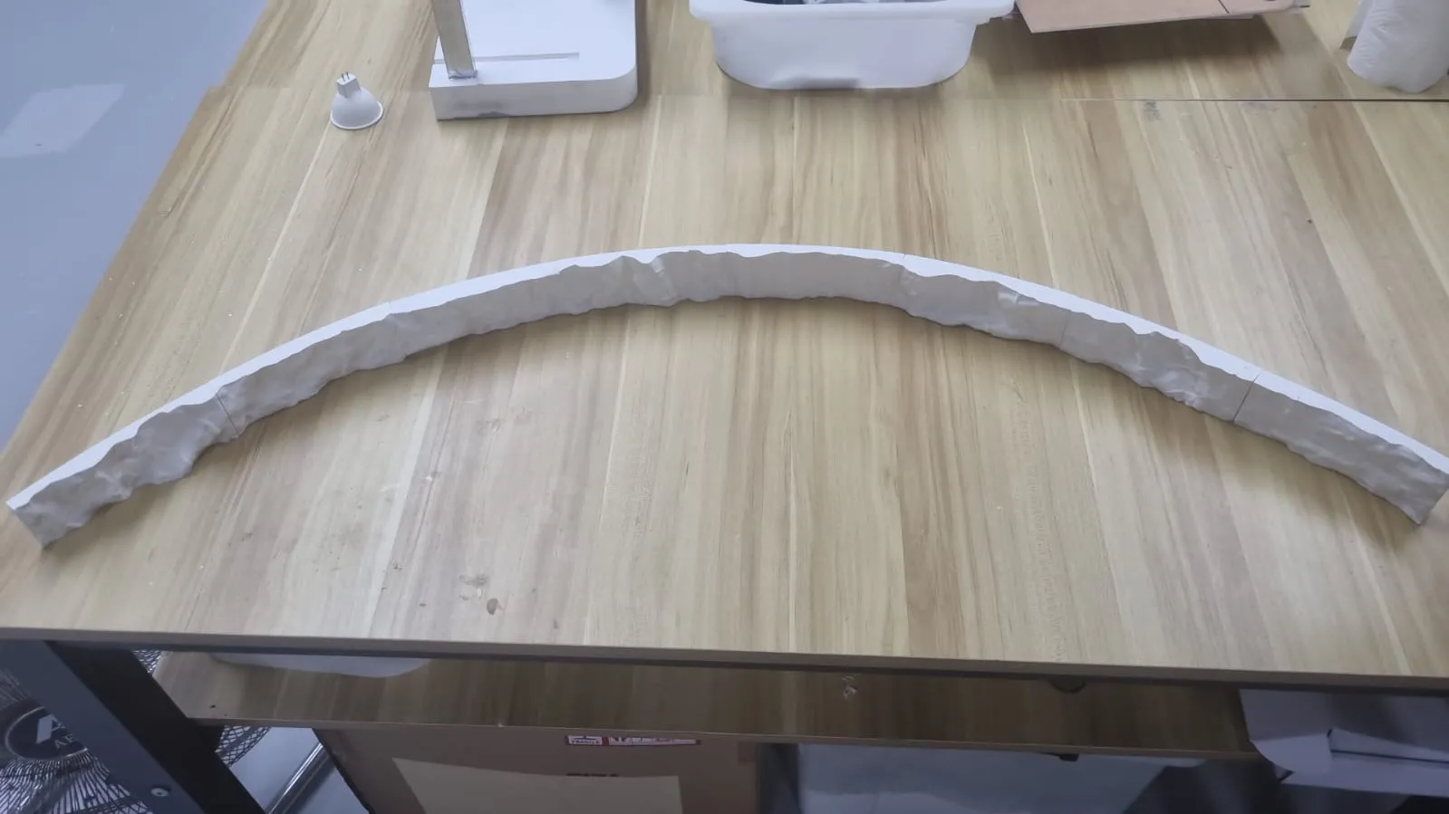 A long white 3D printed strip with a rough rock texture on its face, laid in an arc on a wooden table
