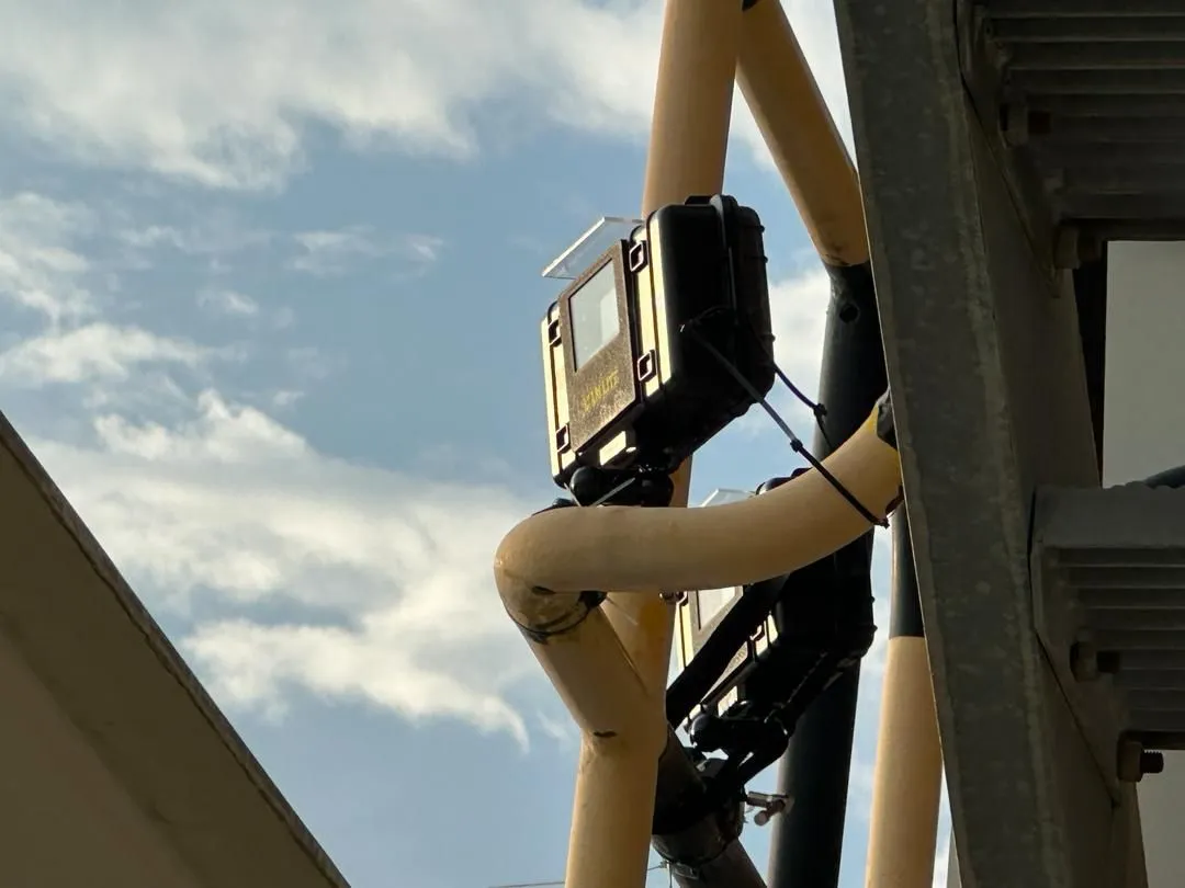 Two XCam Lite housings clamped one above the other to a yellow pipe railing outdoors, against a blue sky