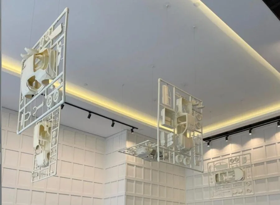 Giant white model car kits, each car and its parts still on the frame, hanging at angles under the lit ceiling of a Toyota showroom café