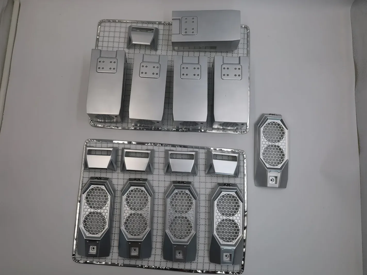 Arc case parts printed in MJF nylon and sprayed silver, laid out on two wire racks: front panels with honeycomb grilles, top caps and rear panels