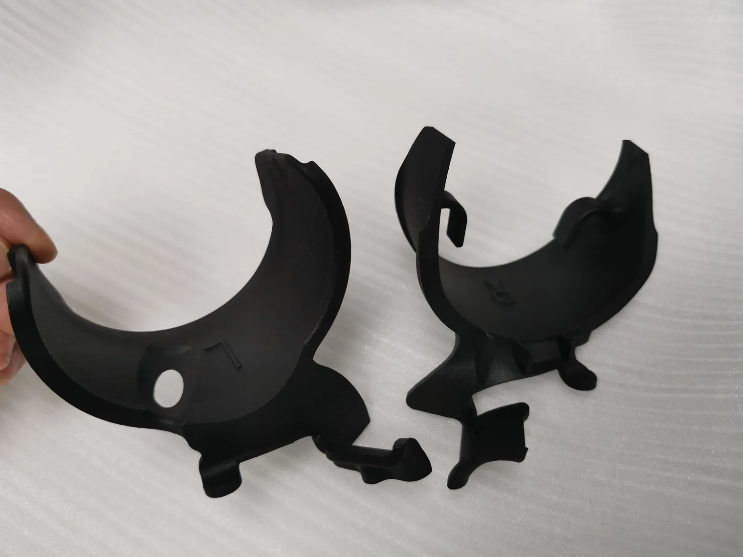 Two black nylon half-shells printed by SLS, one held by its edge, marked L and R, with clips and a fixing hole, showing a matte, slightly grainy surface