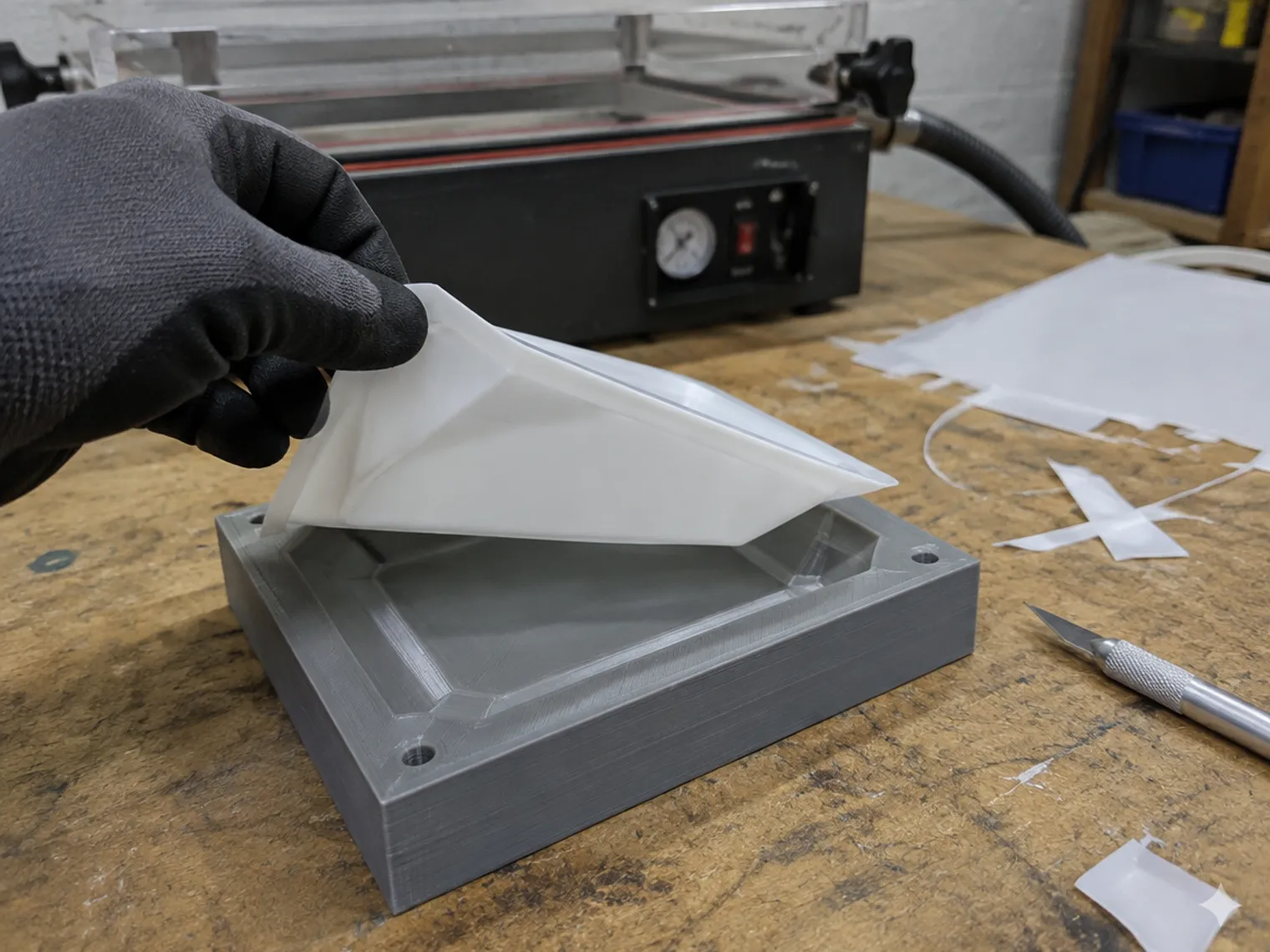 A gloved hand peeling a thin white formed tray off a grey 3D printed mould, the vacuum former behind it