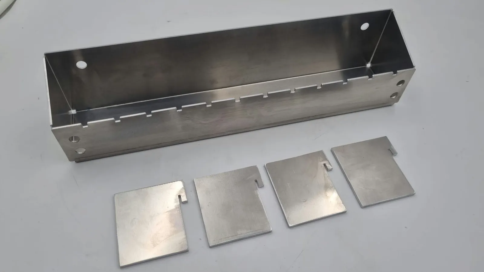 An aluminium tray bent from sheet, with a row of small notches cut along one folded side, and four flat divider plates lying in front of it, each with a notch cut to slot into the tray